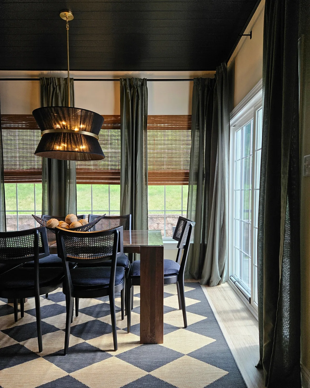 Dining room with a black plank ceiling, black woven drum pendant, cane-back chairs around a walnut table and a navy-and-cream checkerboard rug