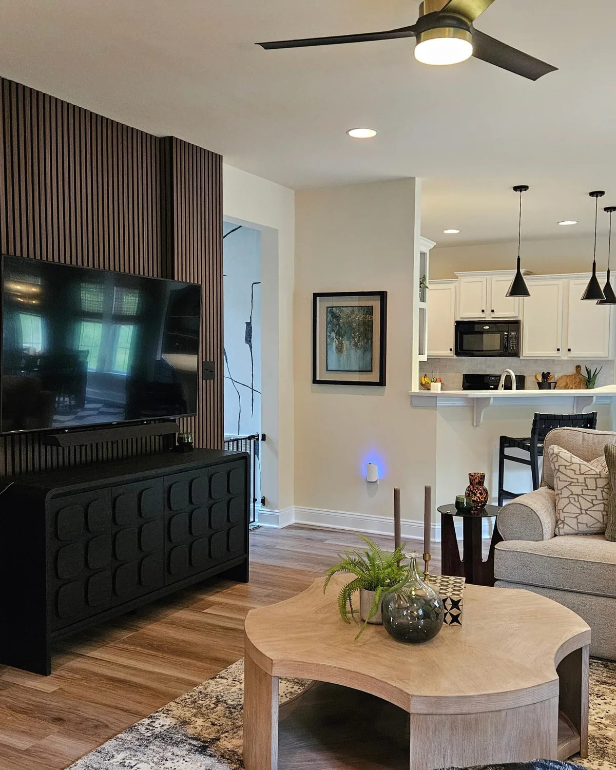 Living room with a vertical wood-slat TV wall, black carved media console and a round wood coffee table, opening to the kitchen