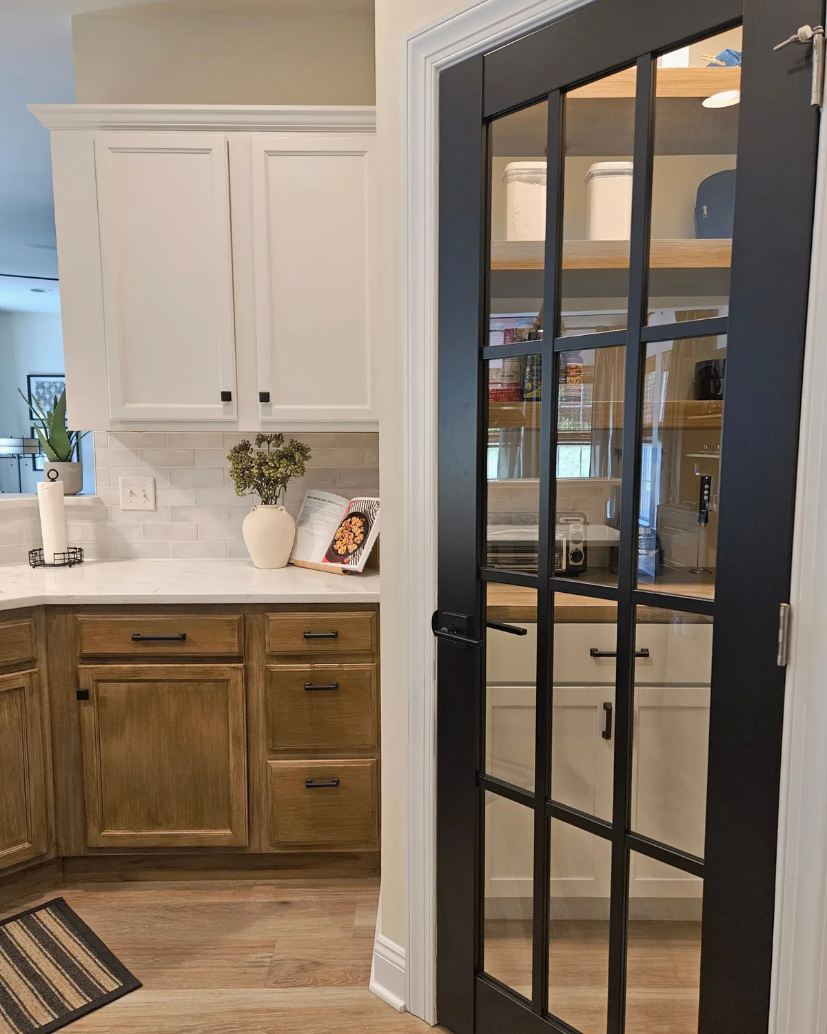 Kitchen seen through a black-framed glass door: white upper cabinets, warm wood lower cabinets and white counters