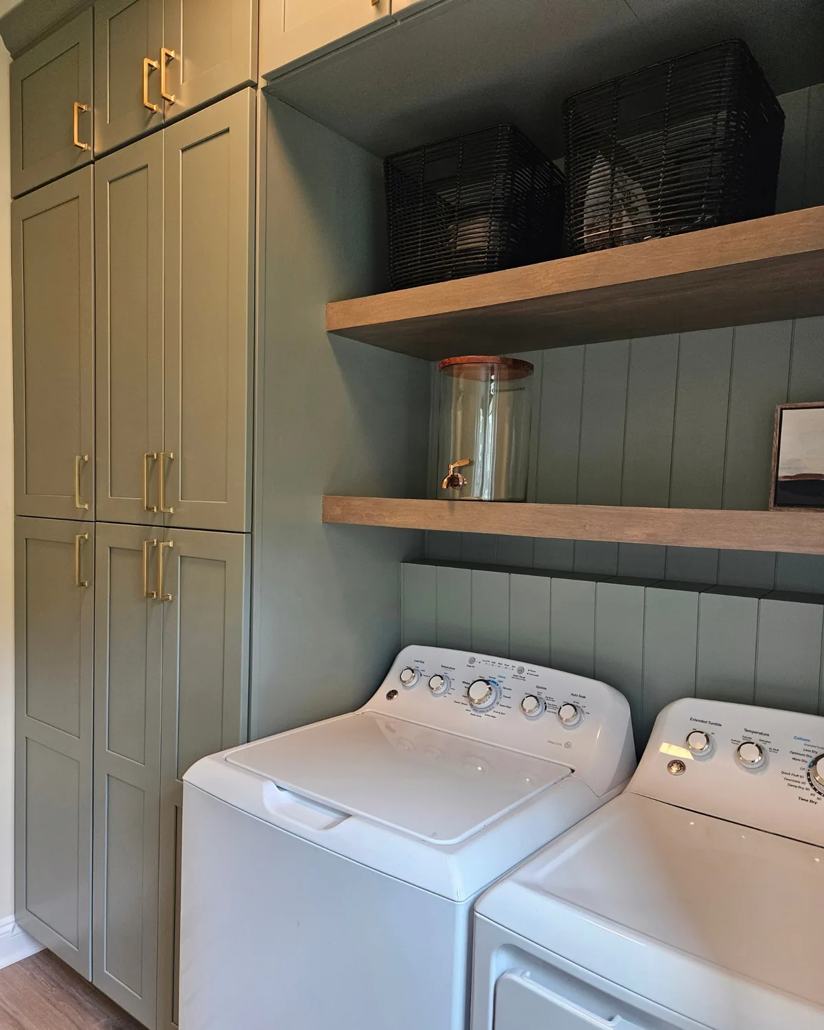 Laundry room with sage green cabinets and beadboard, floating wood shelves and black woven baskets above the washer and dryer