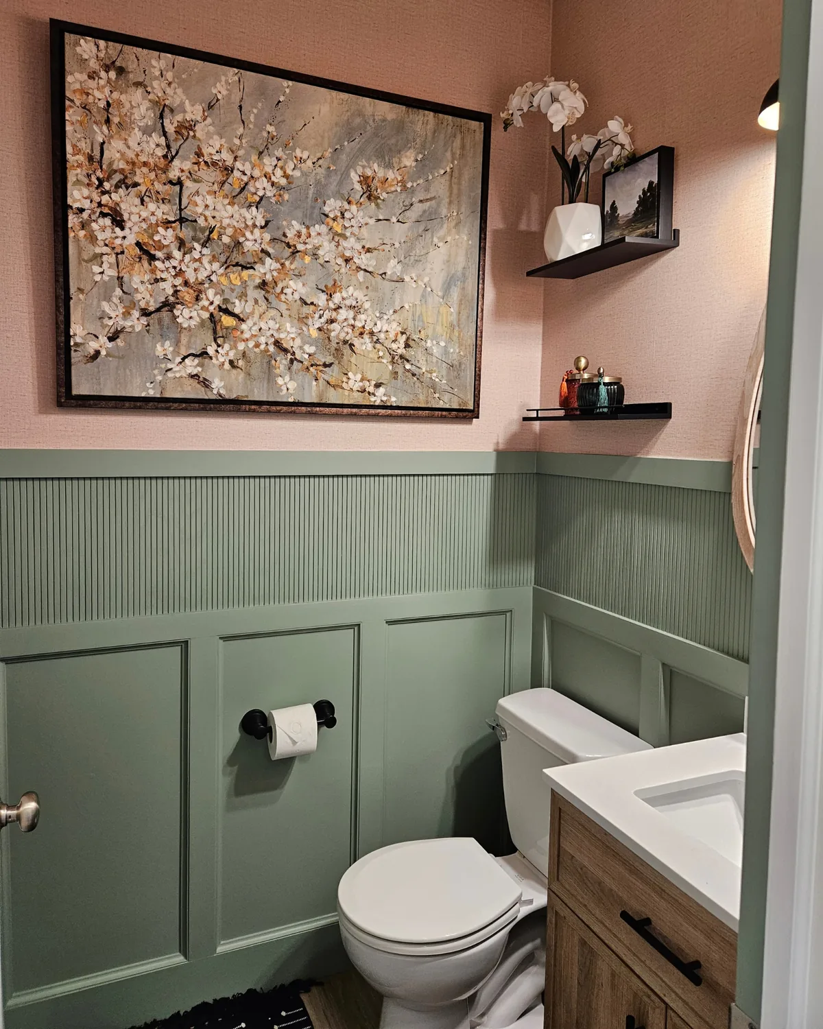 Powder room with blush grasscloth walls above sage green wainscoting, a cherry-blossom painting and a wood vanity