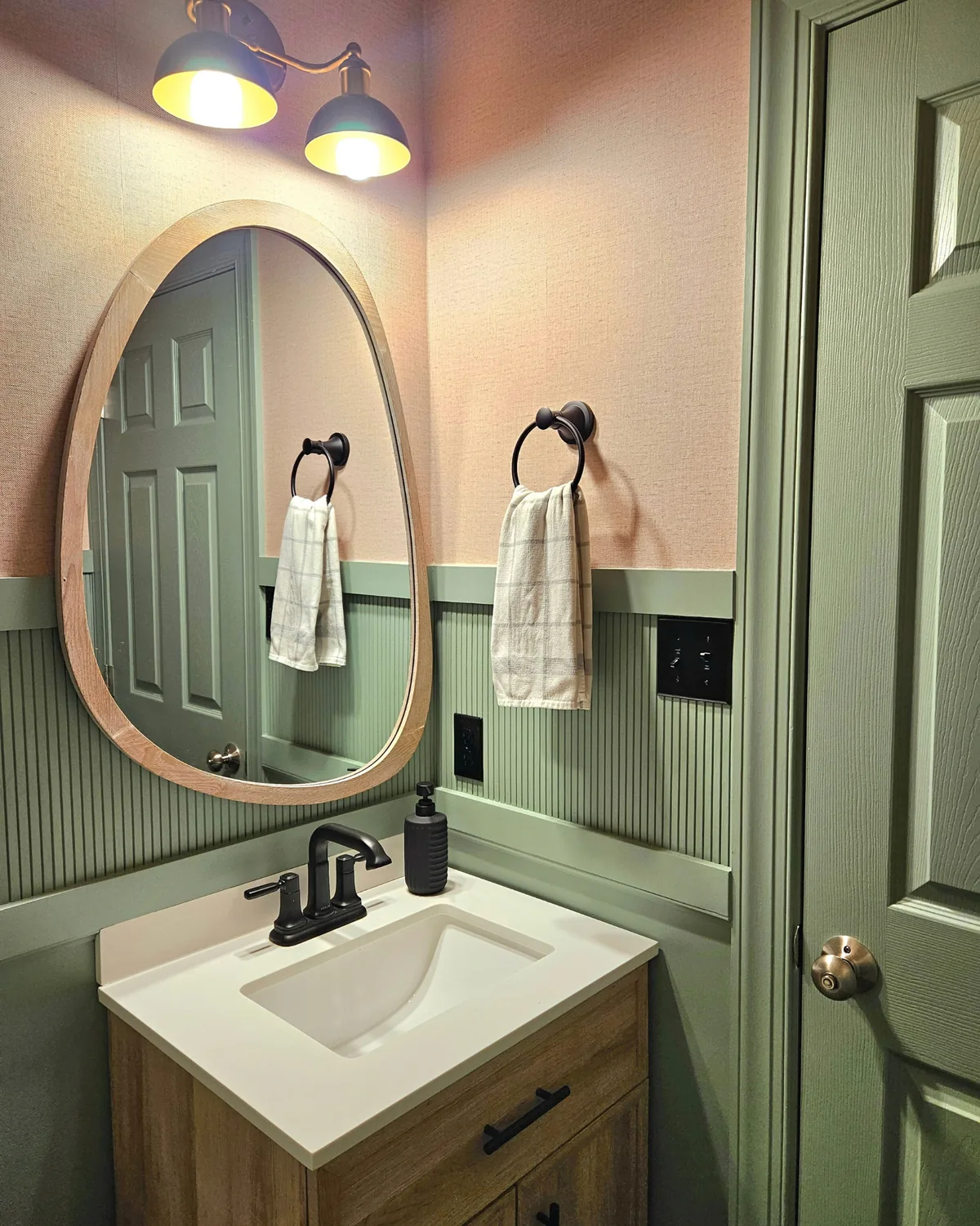 Powder room vanity with an organic oval wood mirror, black faucet and a sage door, lit by a two-light black sconce