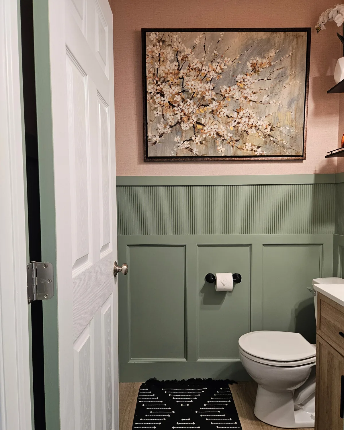 Powder room with a sage door and paneling, blush walls, a cherry-blossom painting and a black-and-white patterned rug