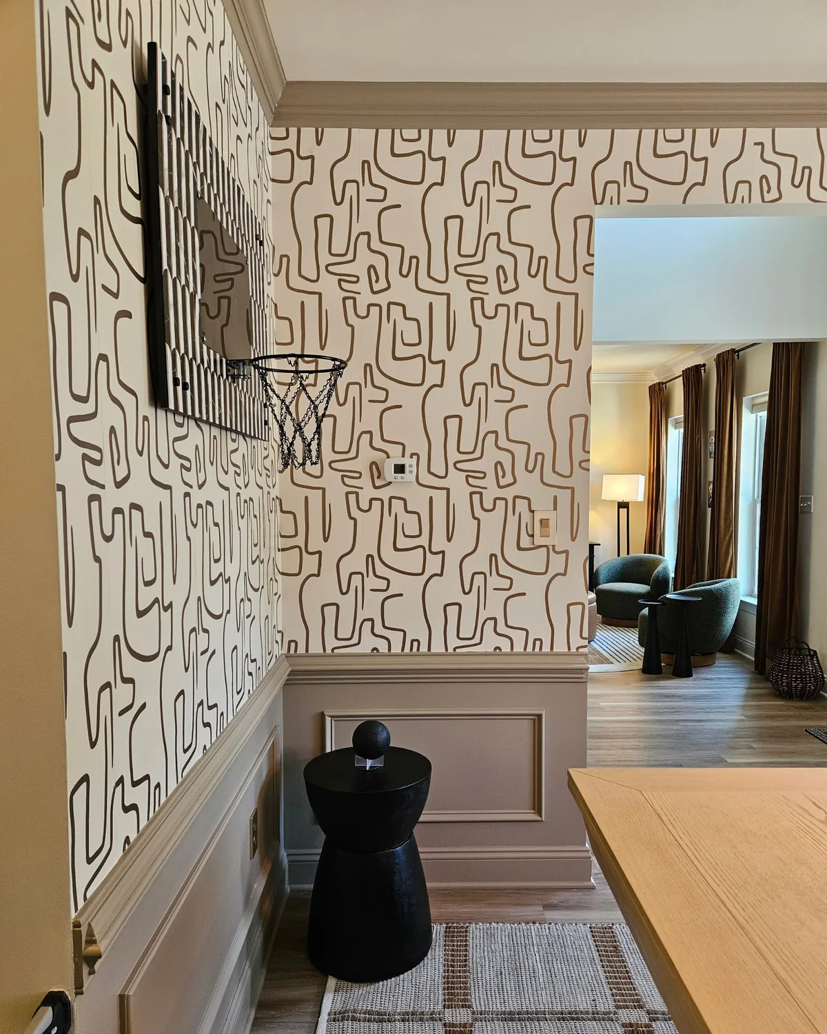Entry with black-and-cream squiggle wallpaper over taupe wainscoting, a black sculptural side table and a view into the living room