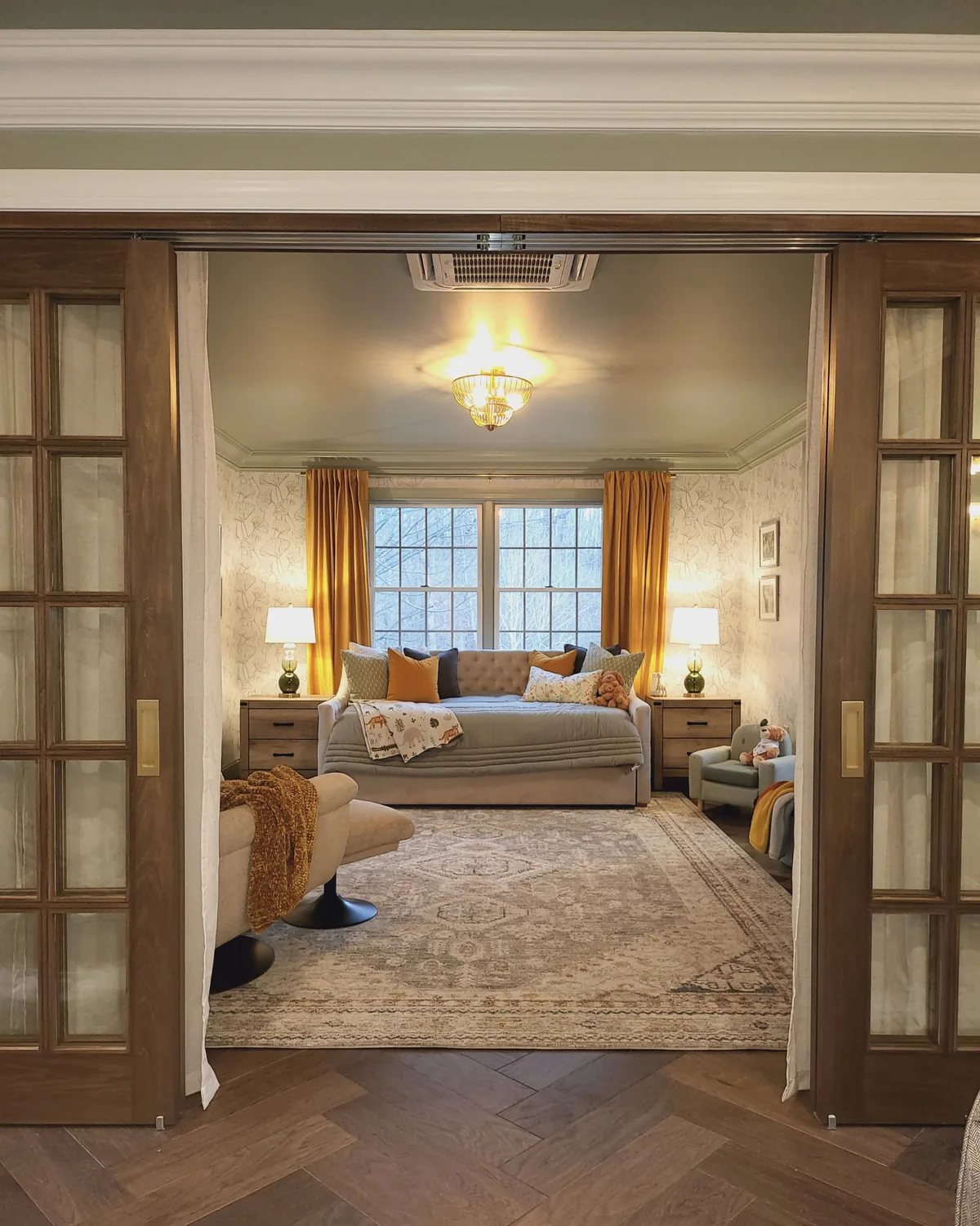 View through wood-framed French doors into a cozy nook with a tufted daybed, amber curtains and botanical wallpaper