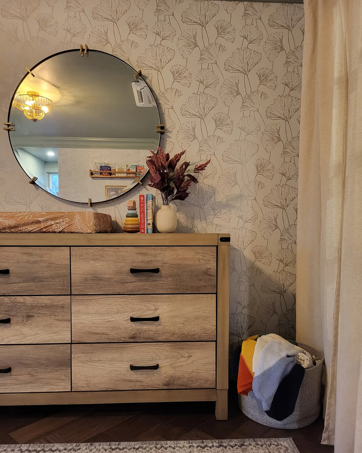 Weathered-oak dresser beneath a round brass mirror against ginkgo-leaf wallpaper, with a basket of throws