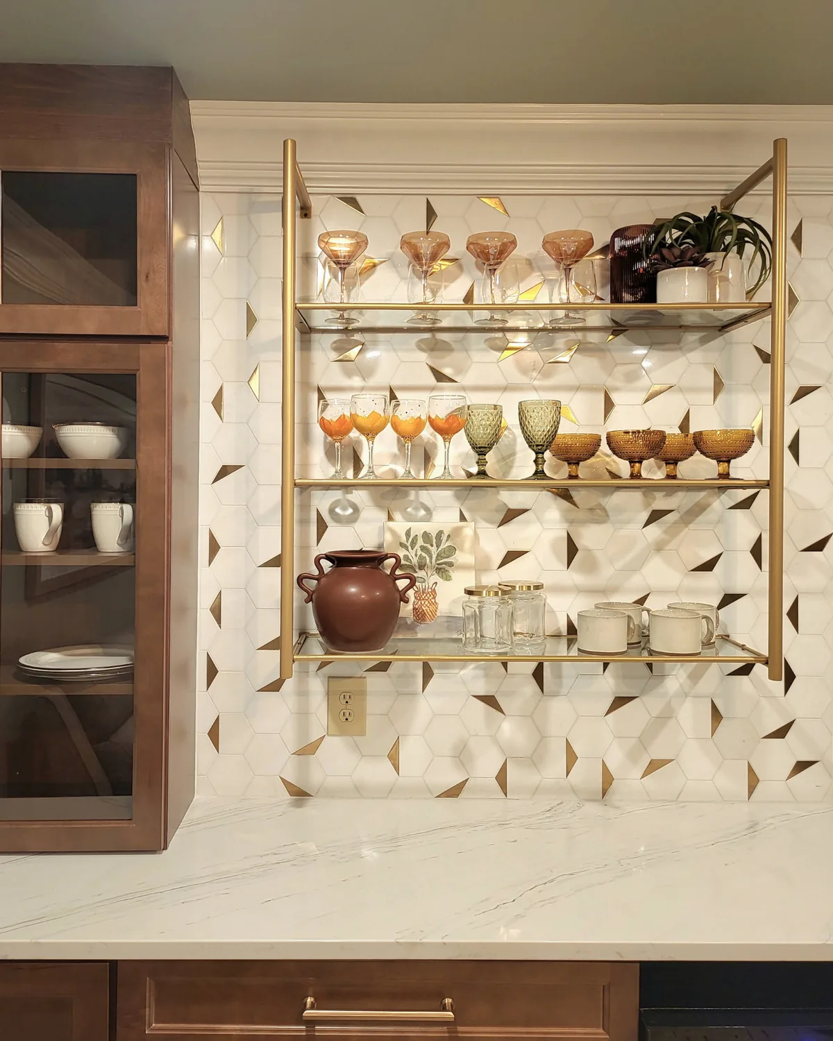 Speakeasy-style bar with brass open shelving of amber coupes and glassware over gold-and-white hexagon tile