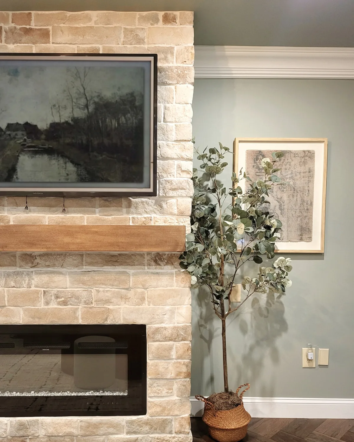 Whitewashed brick fireplace with a reclaimed-wood mantel and a framed-art TV, beside a potted olive tree