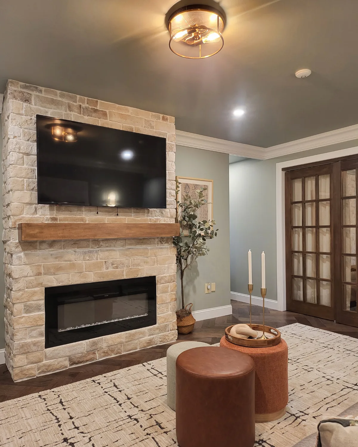 Brick fireplace with a wall-mounted TV, leather ottomans and a plaid rug, with French doors beyond