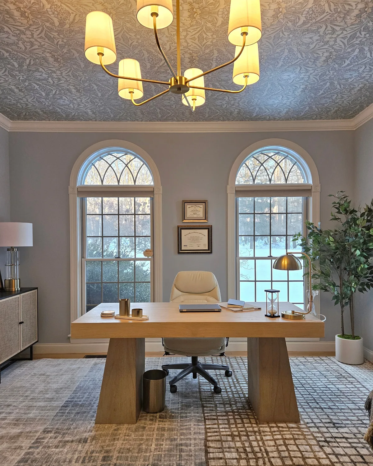 Office with a botanical wallpapered ceiling, a brass chandelier, twin arched windows and a pale oak desk