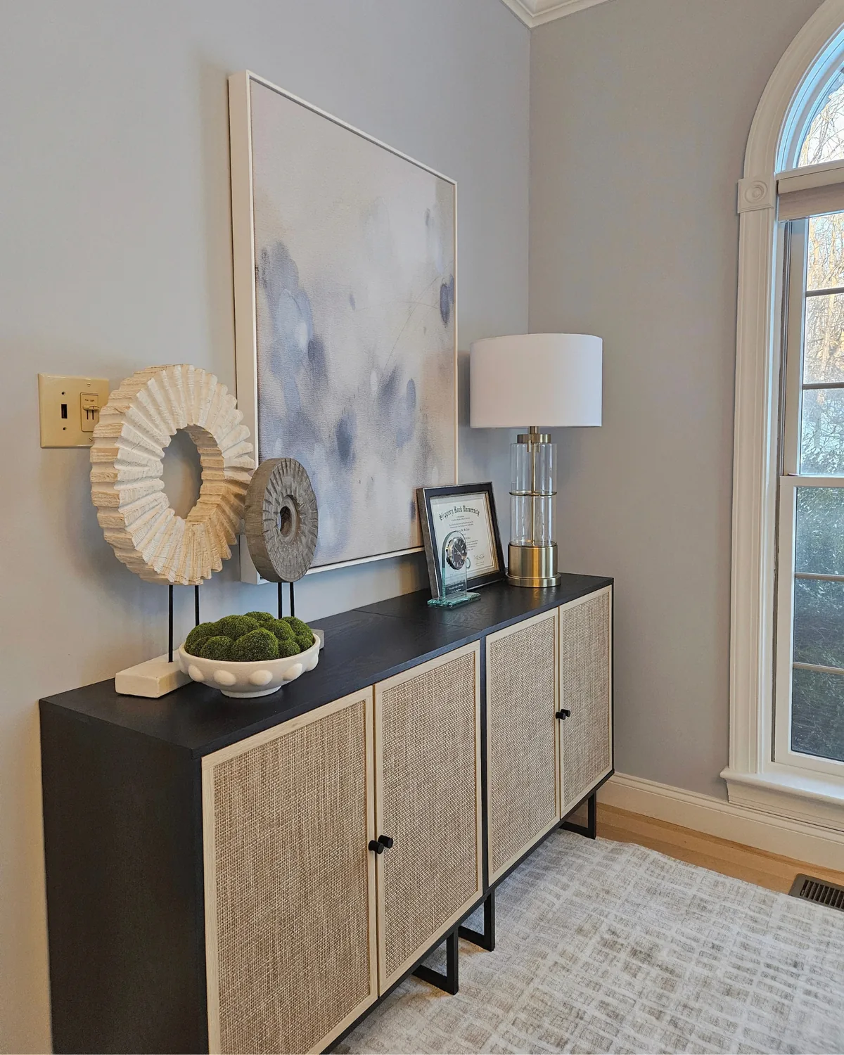 Black-and-cane console styled with sculptural objects, a lamp and soft abstract art beside an arched window