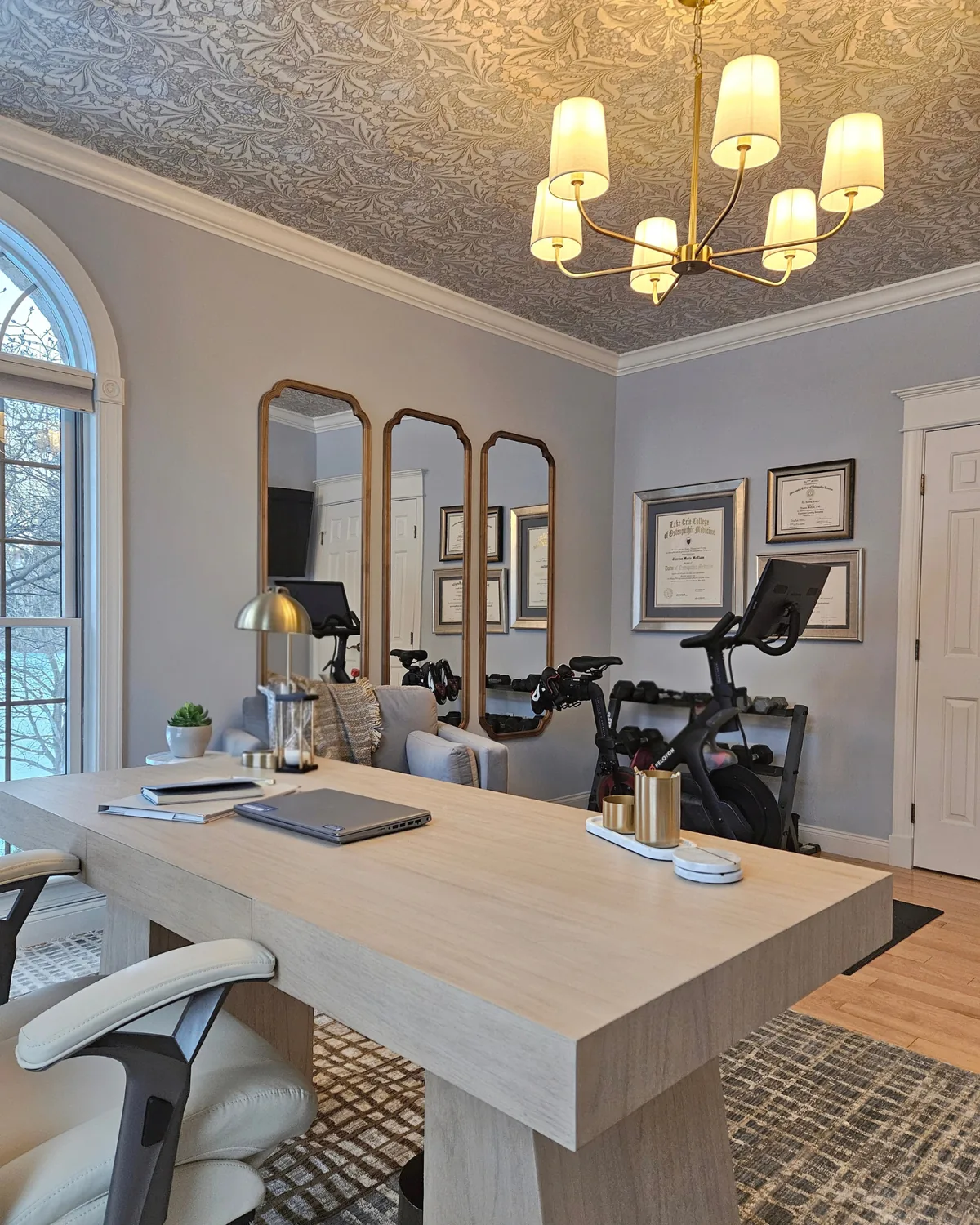 Office and home gym: an oak desk in front, with tall arched brass mirrors, a stationary bike and a dumbbell rack behind