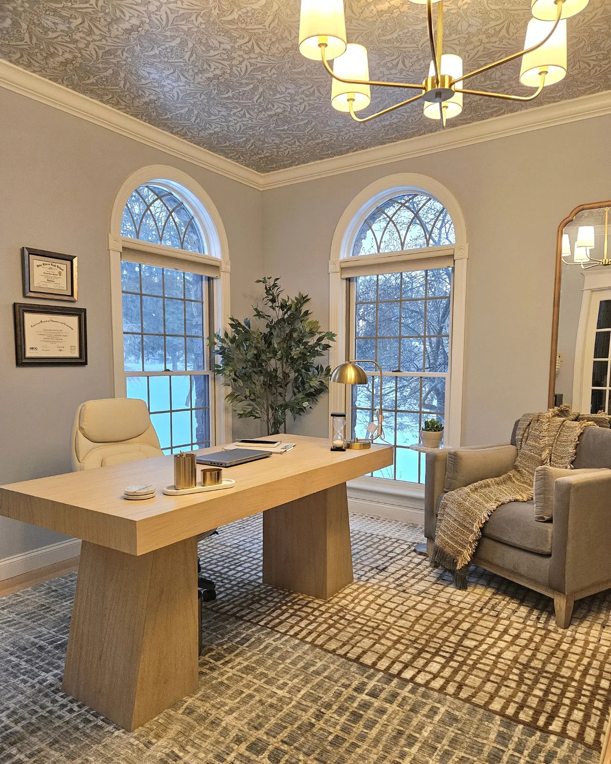 Office with twin arched windows, an oak desk, an armchair with a throw and a wallpapered ceiling