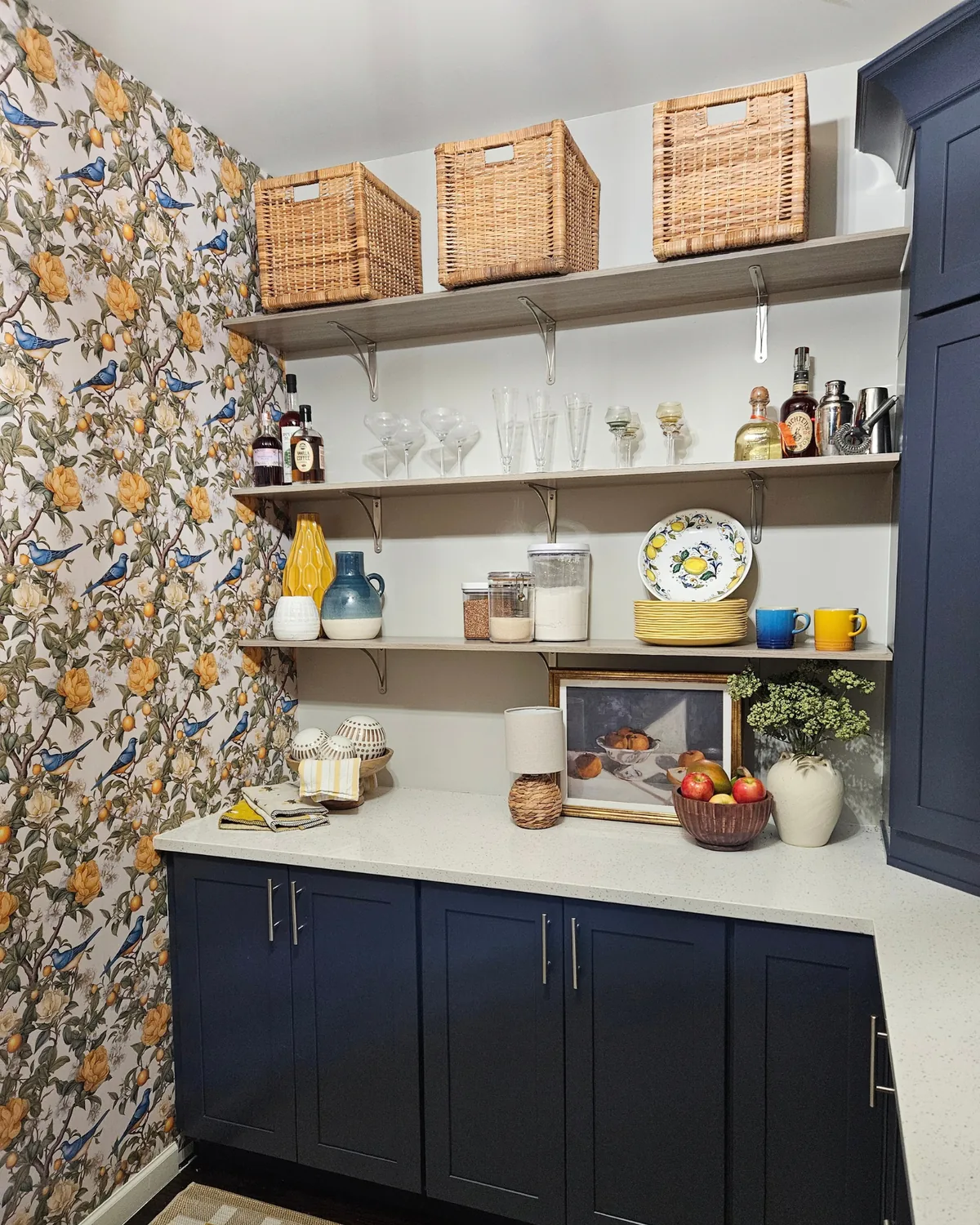 Walk-in pantry with lemon-print wallpaper, open shelves of woven baskets and glassware, navy lower cabinets and a white quartz counter