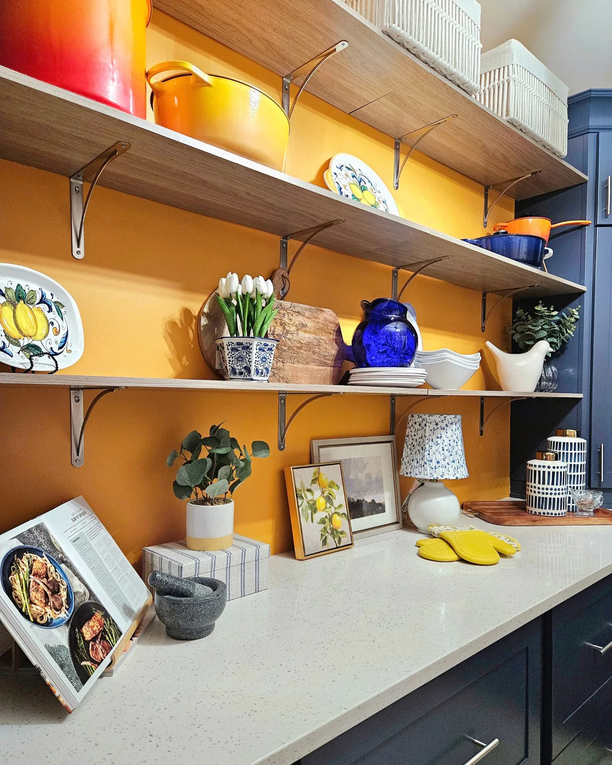 Open wood shelves on a saffron-yellow wall, styled with lemon-patterned plates, cobalt blue vases and orange cookware