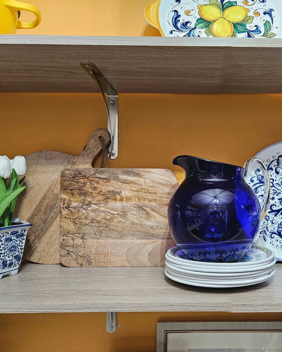 Cobalt blue pitcher, wooden cutting boards and stacked white plates on a wood shelf against a saffron wall