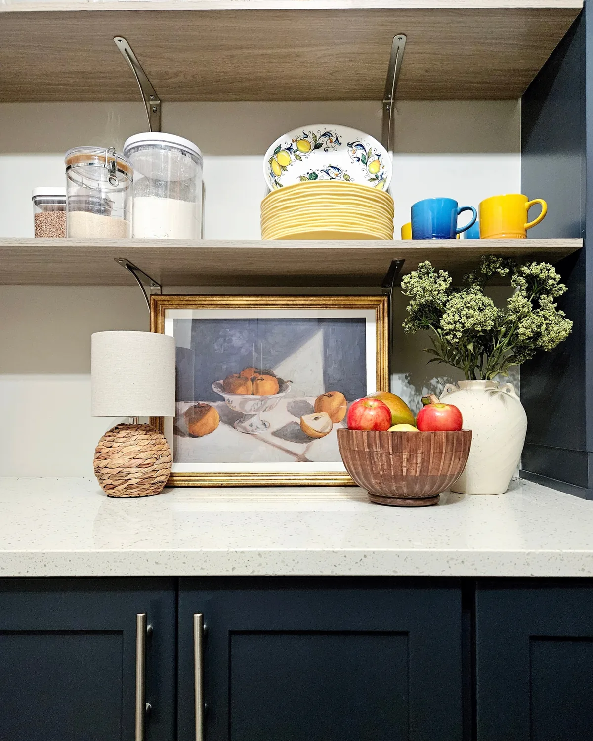 Shelf styled with a still-life painting, a bowl of apples, a white vase of greenery and yellow mugs above navy cabinets