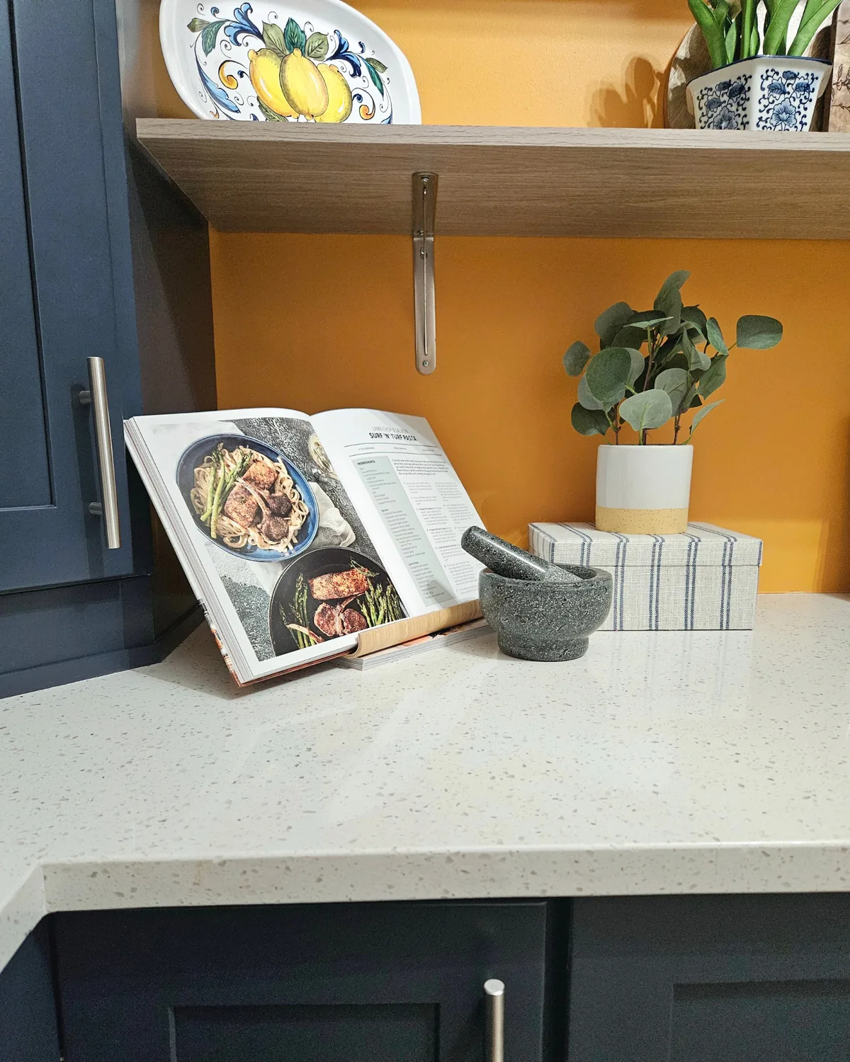 Pantry corner with an open cookbook on a stand, a mortar and pestle and a lemon plate against a saffron wall