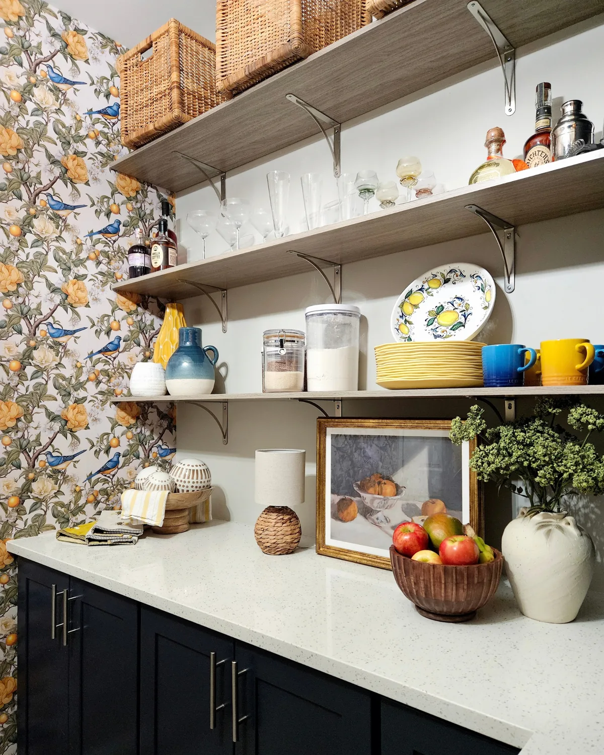 Open shelves of glassware, lemon plates and blue and yellow mugs beside the lemon wallpaper, above a styled quartz counter