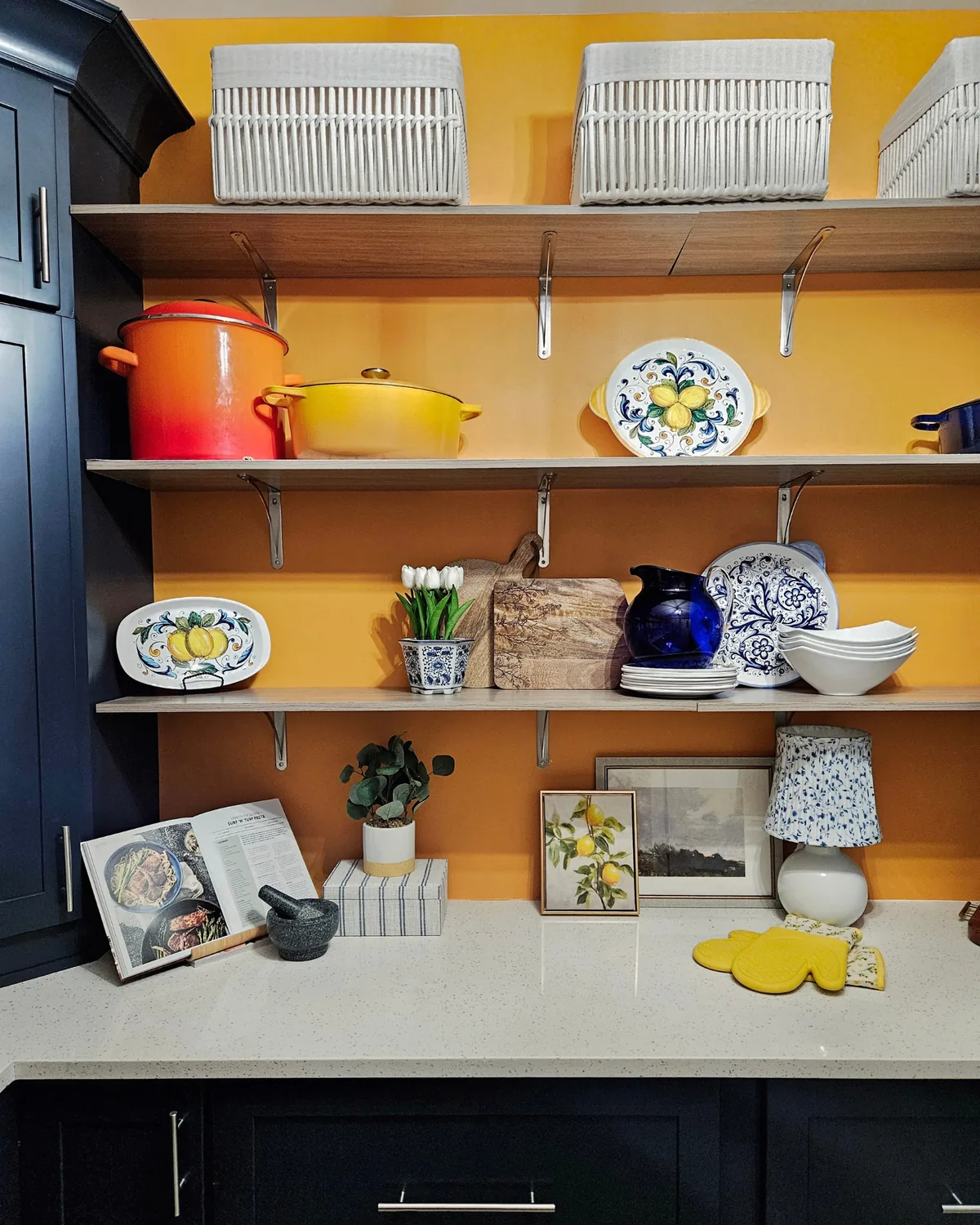 Saffron wall with open shelves of white baskets, orange and yellow cookware, lemon plates and framed art