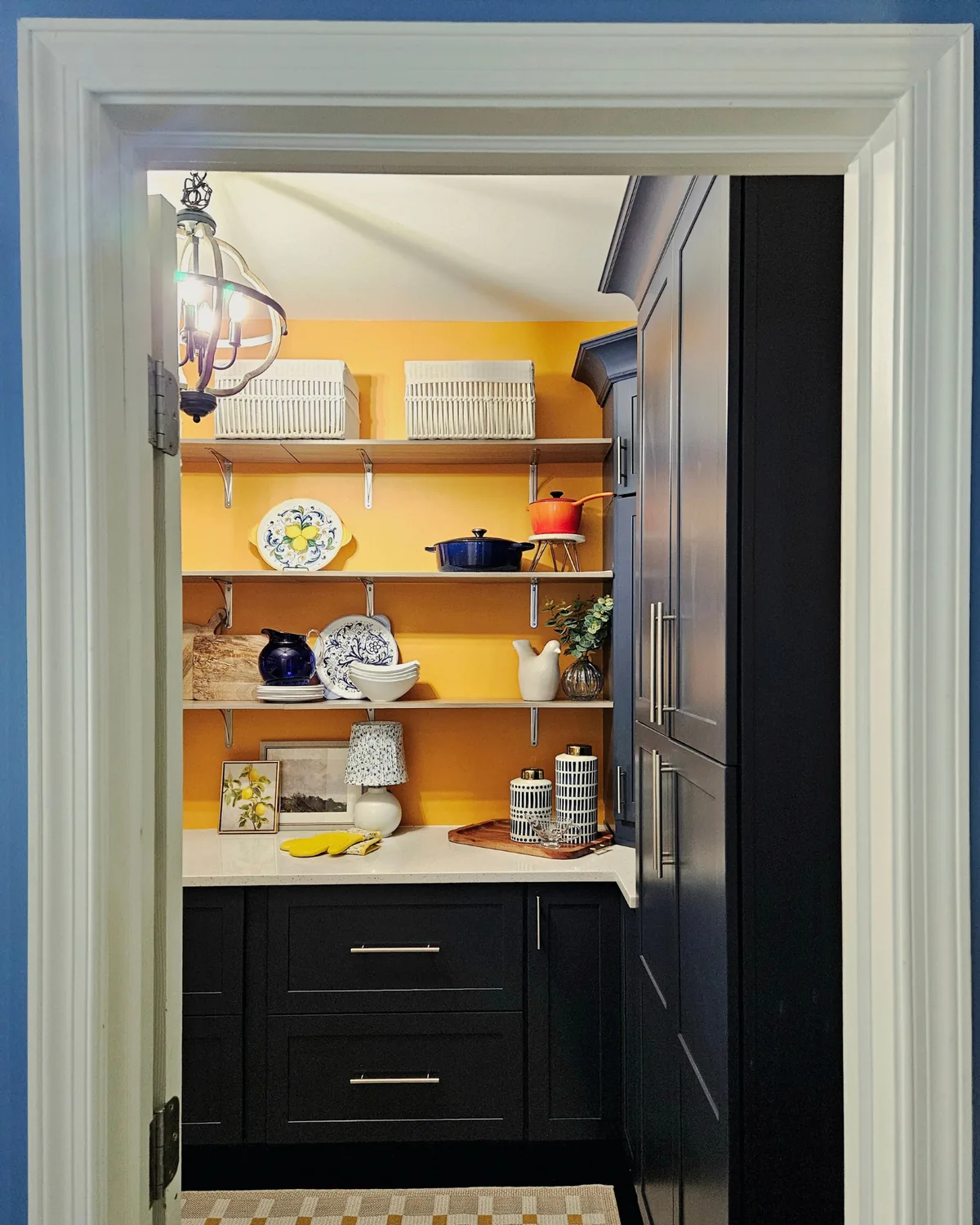 View through the doorway into the pantry: saffron walls, open shelves and navy cabinets beneath a lantern pendant