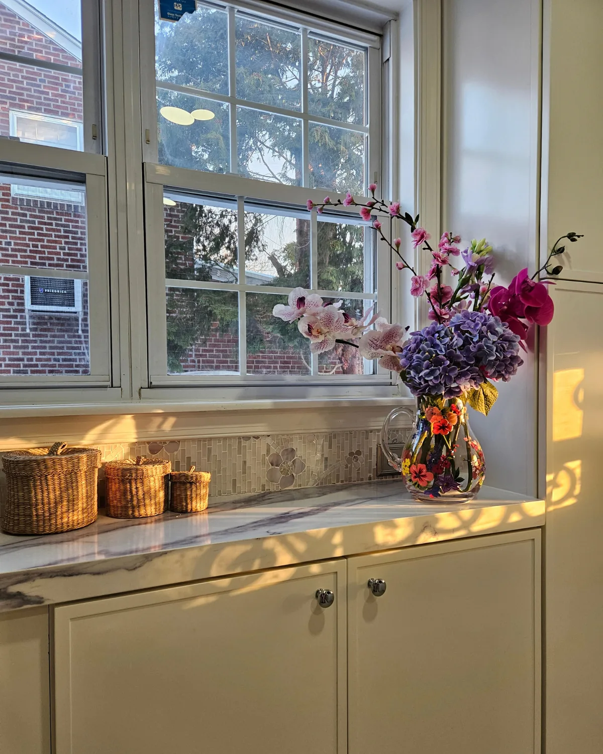 Kitchen window corner with a bouquet of hydrangeas and orchids, woven baskets and lavender-veined marble counters in afternoon sun