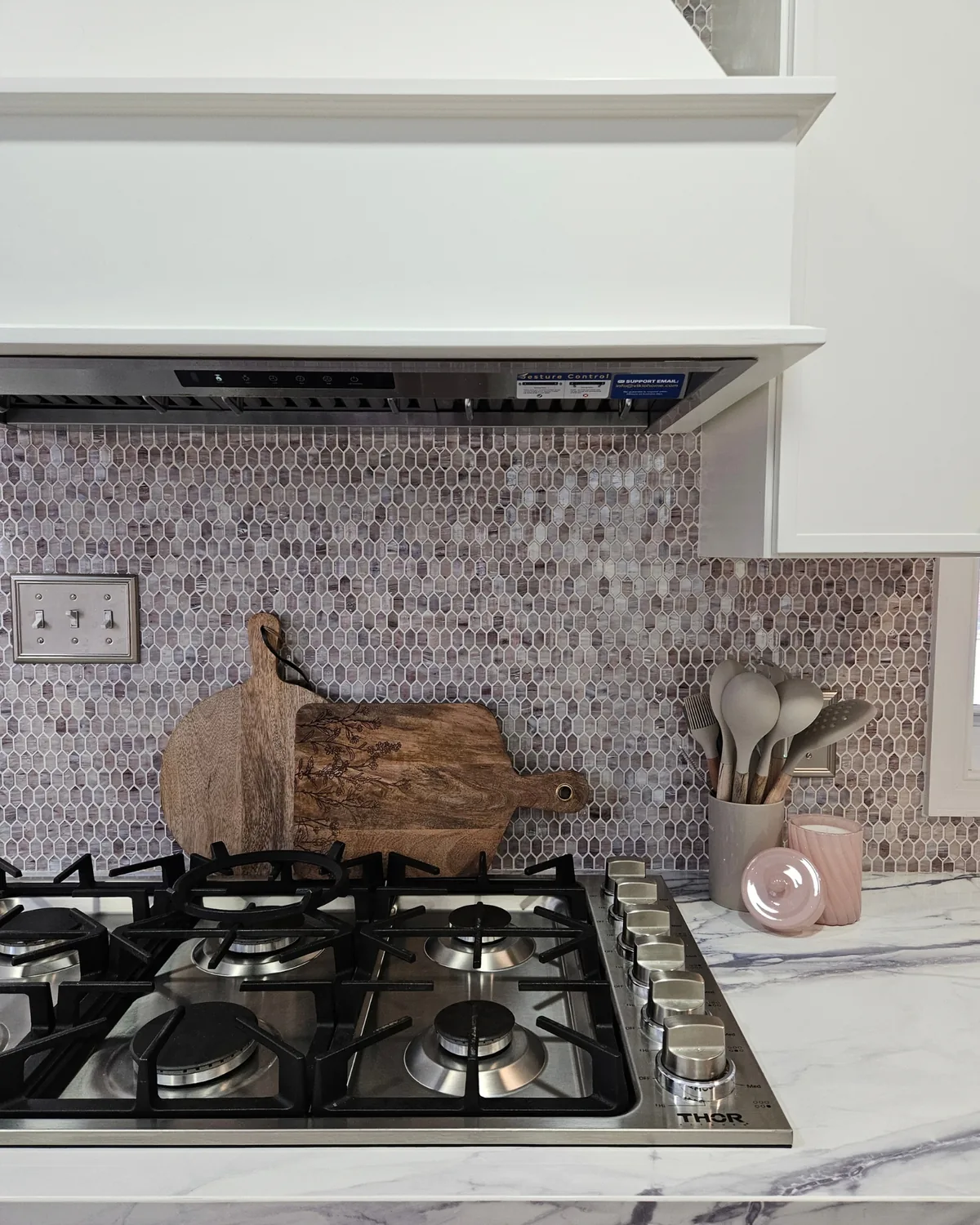 Gas cooktop under a white range hood, with a lavender penny-tile backsplash, wooden cutting boards and blush utensil jars