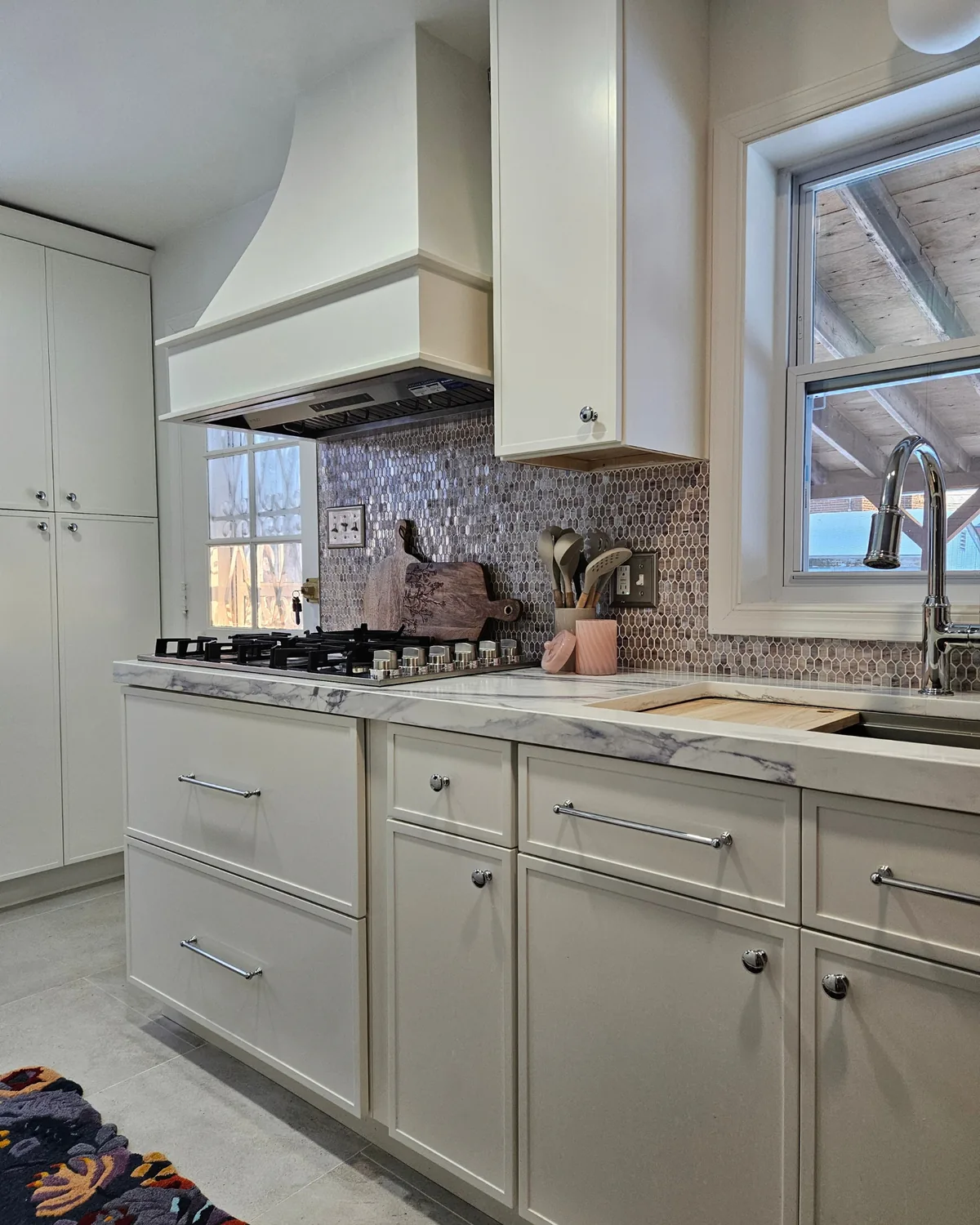 Kitchen run with a white plaster range hood, cream shaker cabinets, lavender mosaic backsplash and a window over the sink