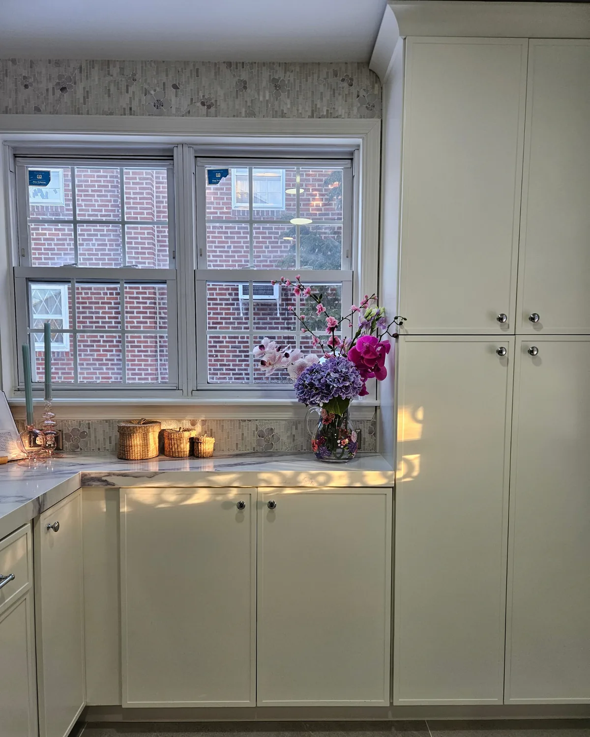 Kitchen corner with tall cream pantry cabinets, a double window and a bouquet of flowers on the marble counter