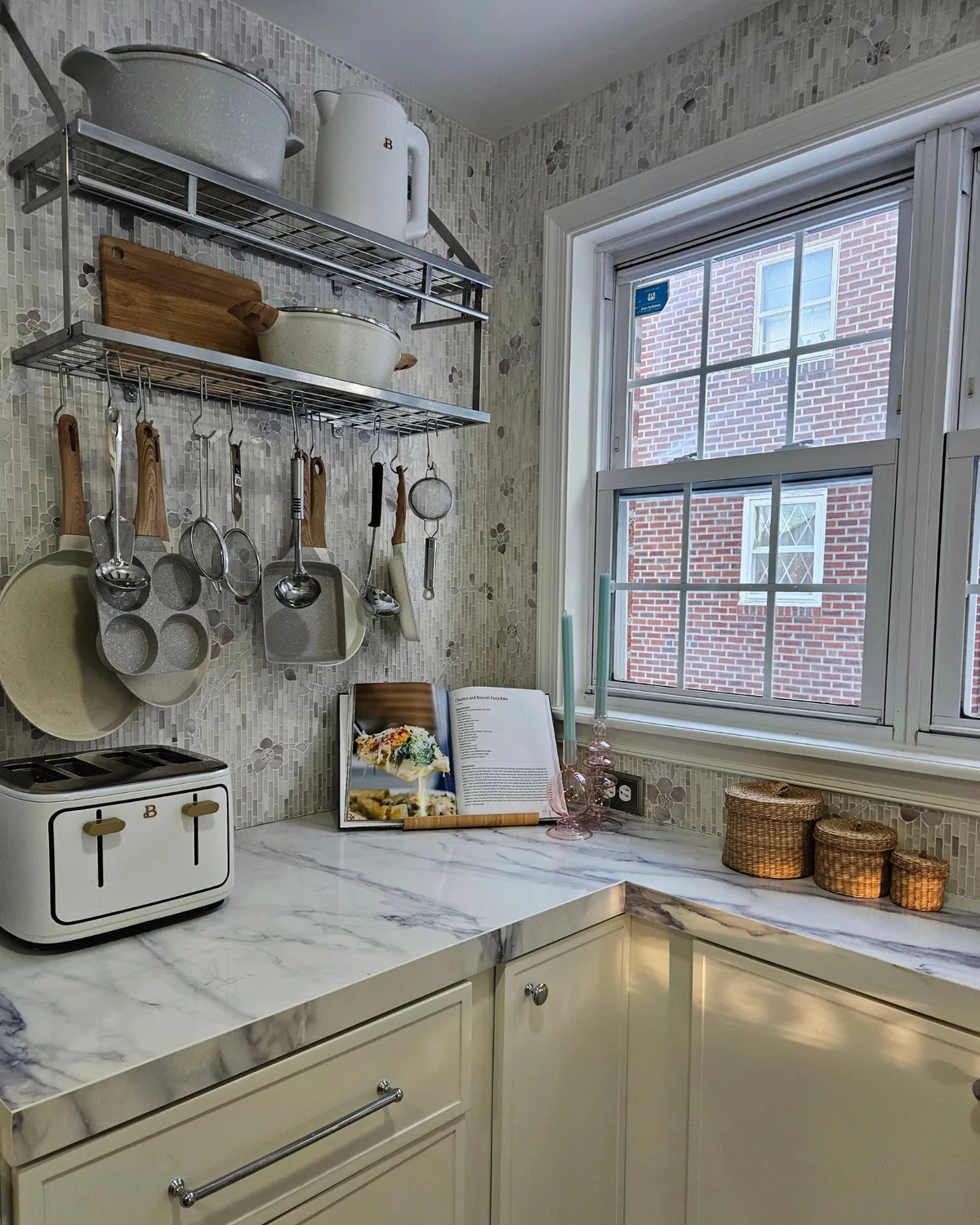 Stainless rail shelf of white cookware and hanging utensils over a lavender stone backsplash, with a cookbook and toaster