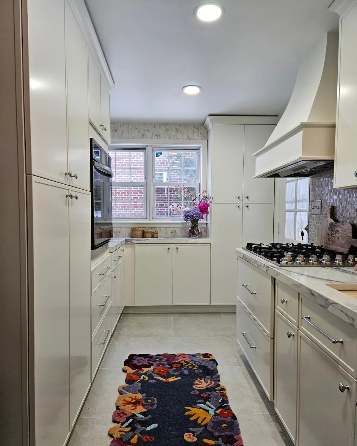 Galley kitchen with tall cream cabinets, a wall oven, the range hood and a floral runner leading to the window