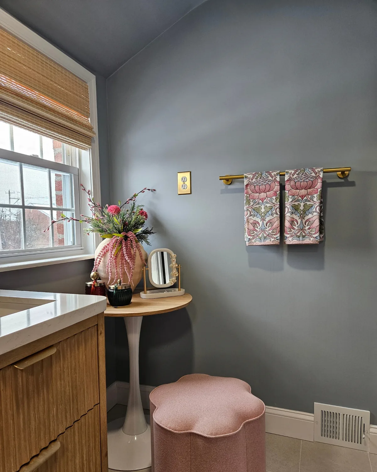 Blue-gray bathroom with a brass towel bar of floral towels, a tulip table and a pink flower stool beside a wood vanity