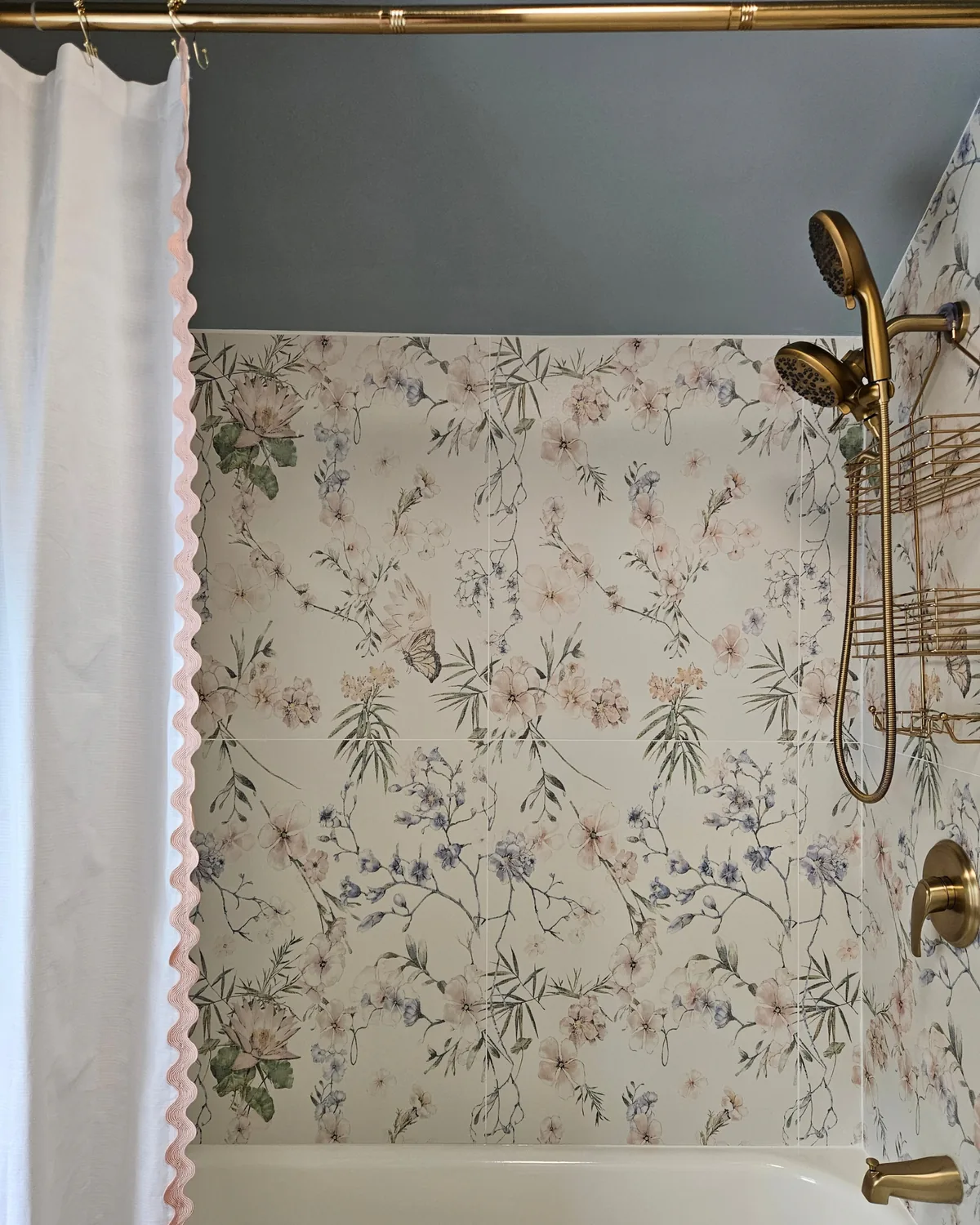 Tub with floral wildflower tile, a brass handheld shower and a white curtain trimmed in pink rickrack
