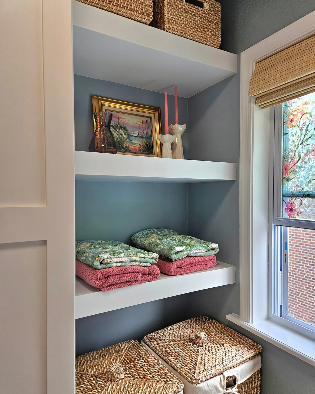 Built-in open shelves with woven baskets, floral towels, pink candles and a framed painting beside a window