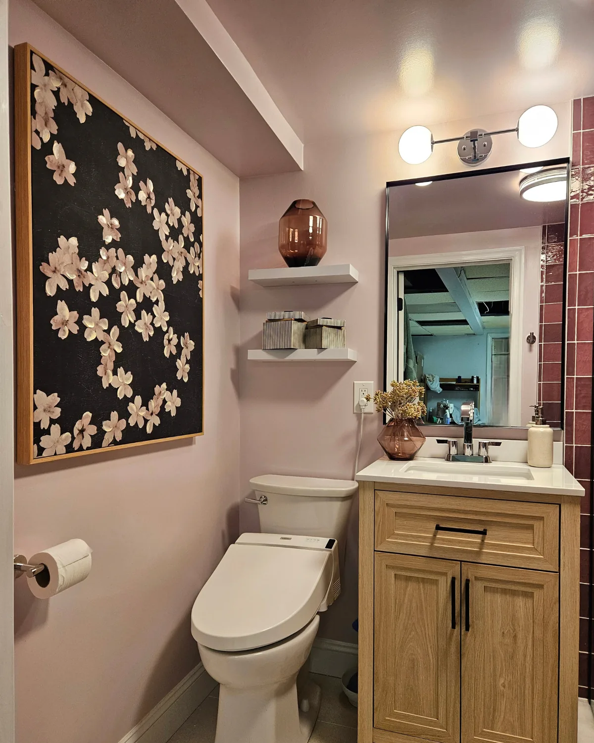 Blush bathroom with cherry-blossom art, a light wood vanity, globe sconce and berry tile at the shower edge