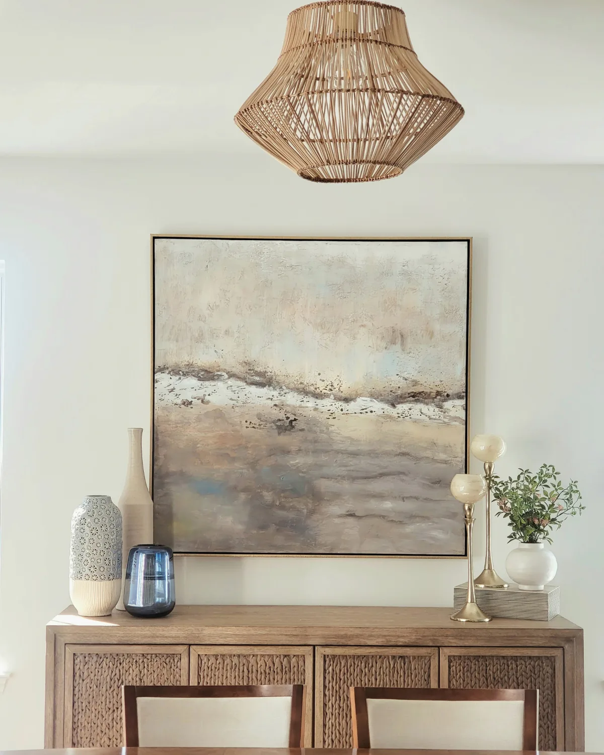 Dining sideboard styled with a sea-toned abstract painting, brass candlesticks and greenery under a rattan pendant