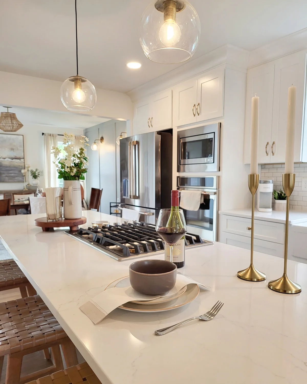 Quartz island set for dinner with a gas cooktop, brass candlesticks and wine, with white cabinets and stainless appliances behind