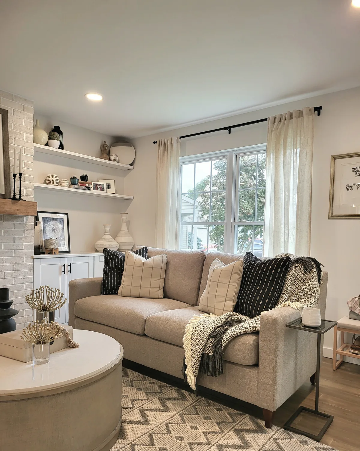 Living room with a greige sofa, black-and-cream pillows, a round ottoman and floating shelves beside a white brick fireplace