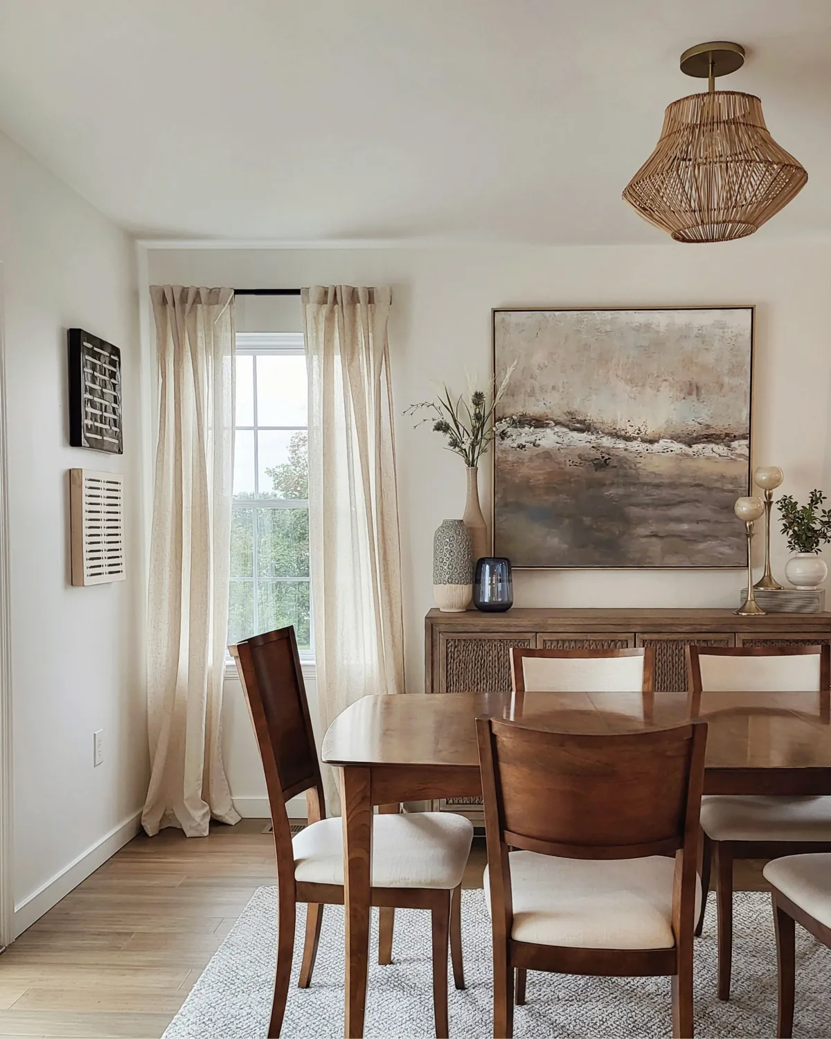 Dining room with linen curtains, a rattan pendant, a wood dining set and the sea-toned painting over the sideboard