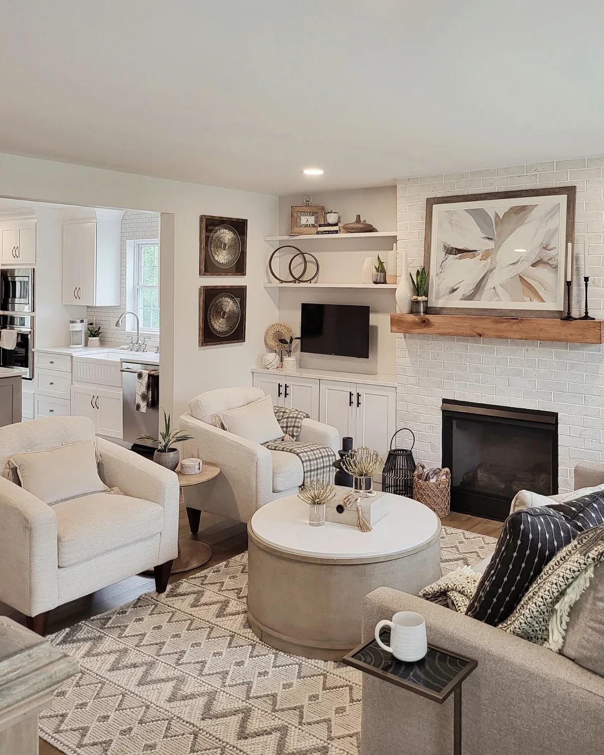 Living room with a white brick fireplace and wood mantel, a round ottoman, cream armchairs and a chevron rug, with the kitchen beyond
