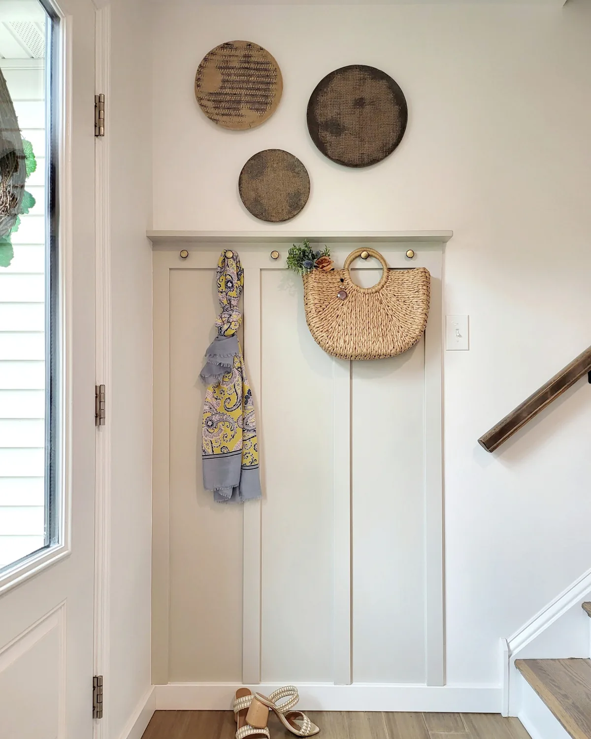 Entry wall with board-and-batten paneling, brass hooks holding a scarf and straw bag, and round woven wall art