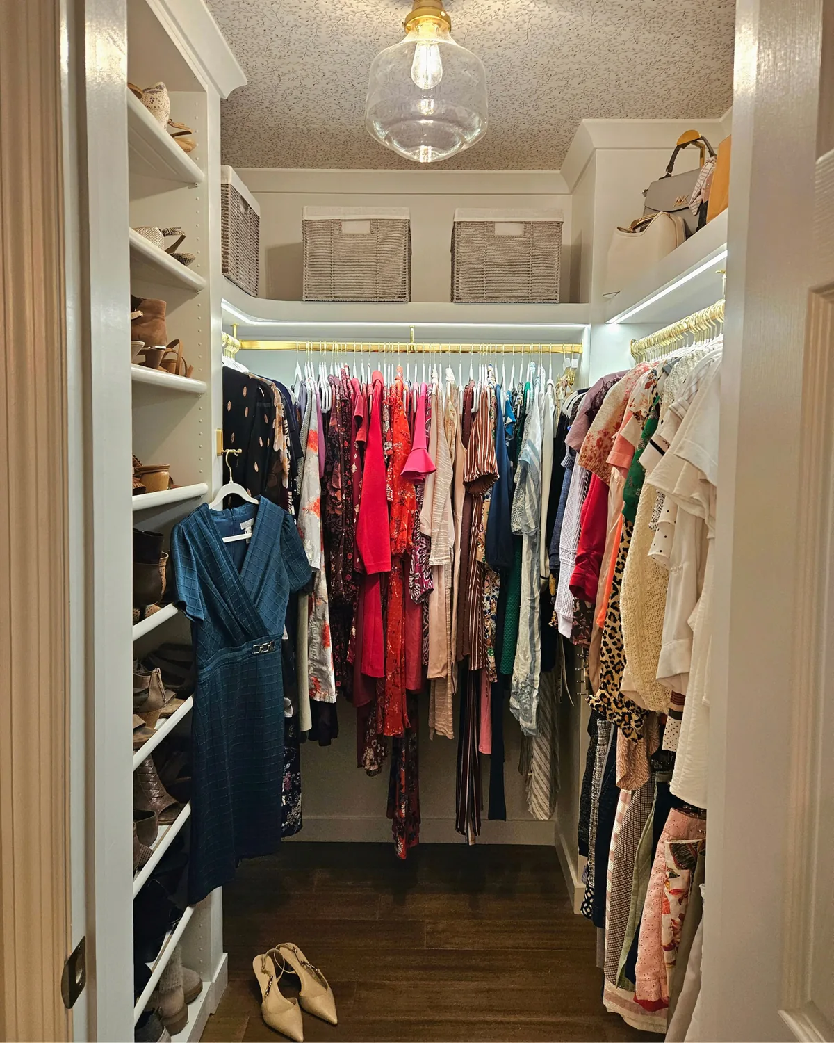 Primary closet in a U-shape with brass hanging rods, a glass pendant and shoe shelving
